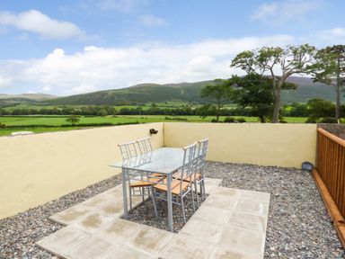 An outdoor patio with a glass table and four chairs surrounded by gravel and low yellow walls with a view of green fields and hills at Fell View Cottage in Bootle near Millom