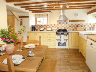A kitchen with wooden beams yellow cabinets a wooden dining table with pink flowers at Fell View Cottage in Bootle near Millom