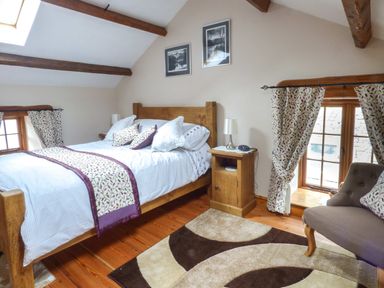 A bedroom with a wooden bed a bedside table a lamp a chair and two windows with patterned curtains at Fell View Cottage Bootle near Millom