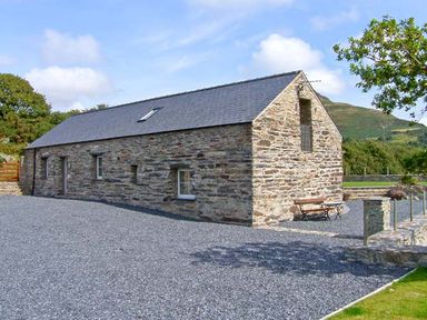 A stone barn with a slate roof and gravel driveway surrounded by trees and hills at Garth Morthin The Barn in Porthmadog