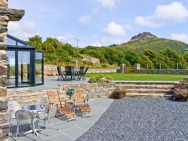 An outdoor patio area with metal and wooden chairs tables stone walls and green grass with a hill in the background at Garth Morthin The Barn in Porthmadog