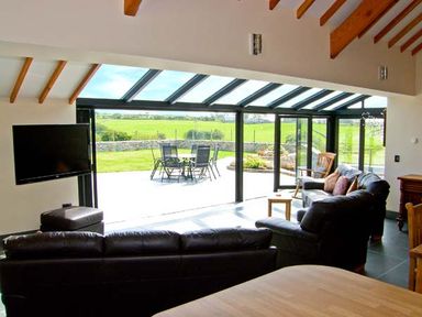 A living room with black leather sofas a wall-mounted TV and large glass doors opening to a patio with outdoor furniture at Garth Morthin The Barn in Porthmadog