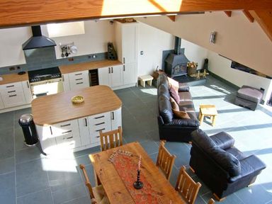 A combined kitchen and living area with wooden dining table and chairs a kitchen island cooking area and black leather sofas at Garth Morthin The Barn in Porthmadog