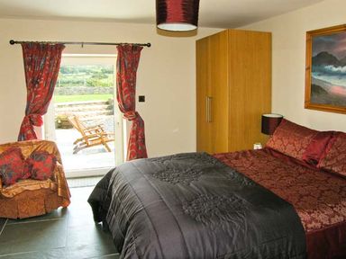 A bedroom with a bed covered in red and black bedding a wooden wardrobe an armchair with cushions red patterned curtains and a glass door leading outside at Garth Morthin The Barn in Porthmadog