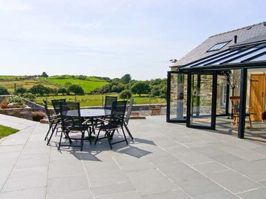 An outdoor patio area with a glass table and chairs next to a stone building with glass doors overlooking green fields at Garth Morthin The Barn in Porthmadog