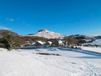 A snowy rural landscape with stone buildings near trees and a hill in the background at Garth Morthin The Barn in Porthmadog