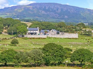 A stone farmhouse and buildings surrounded by a stone wall in a green field with trees and sheep with hills in the background at Garth Morthin The Barn in Porthmadog