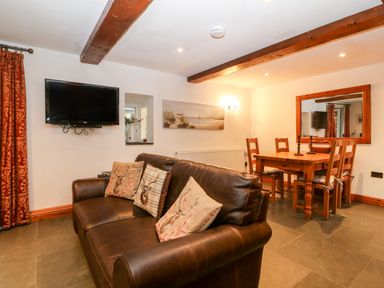 A living room with a brown leather sofa and cushions a wall mounted TV a wooden dining table with chairs and a large wall mirror at Garth Morthin The Stables in Porthmadog