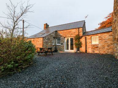 An exterior view of a stone cottage with a slate roof a wooden picnic table and gravel yard at Garth Morthin The Stables in Porthmadog
