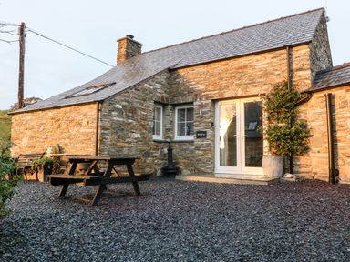 An outdoor patio area with a wooden picnic table bench and stone cottage with glass double doors and plants at Garth Morthin The Stables in Porthmadog