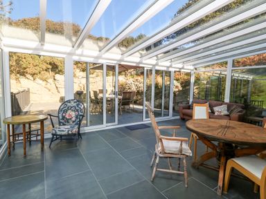 A glass conservatory with a round wooden table and chairs and a floral armchair at Garth Morthin The Stables in Porthmadog