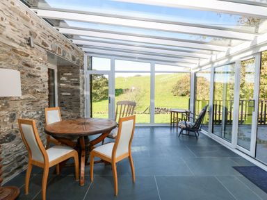 A sunroom with a round wooden table and four chairs a rocking chair and glass walls showing a green field at Garth Morthin The Stables in Porthmadog