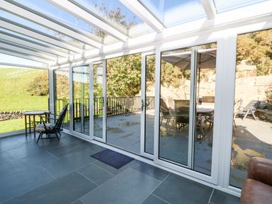 A sunroom with glass walls and ceiling featuring a chair and table at Garth Morthin The Stables in Porthmadog