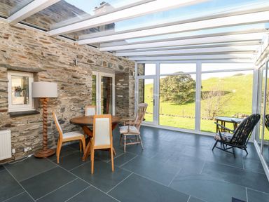 A sunroom with a round wooden table and four chairs stone wall and large windows overlooking green fields at Garth Morthin The Stables in Porthmadog