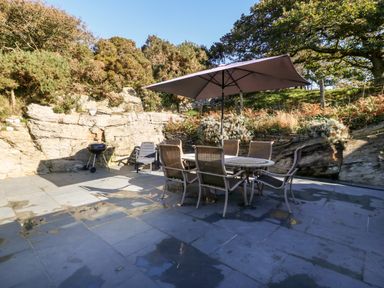 An outdoor patio with a round table, chairs, a large umbrella, and a charcoal grill surrounded by natural rock and plants at Garth Morthin The Stables in Porthmadog