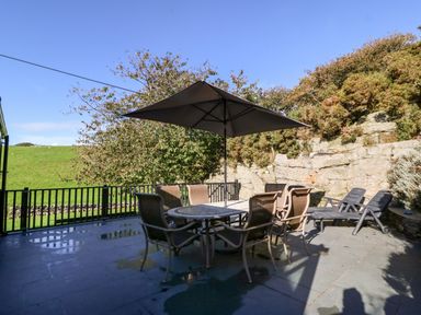 An outdoor patio with a table and six chairs under an umbrella and two lounge chairs beside a stone wall at Garth Morthin The Stables in Porthmadog