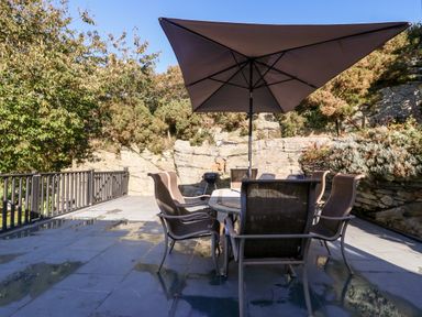 An outdoor patio area with a table and six chairs under a large umbrella and a small charcoal grill near a stone wall at Garth Morthin The Stables in Porthmadog