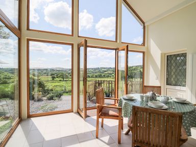 A dining area with a table and chairs next to large windows and doors opening to a garden with a view of fields at Llety'r Wennol in Ffarmers near Lampeter