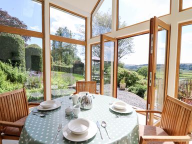 A dining area with a round table set with dishes and cutlery surrounded by wooden chairs and glass doors opening to a garden at Llety'r Wennol Ffarmers near Lampeter