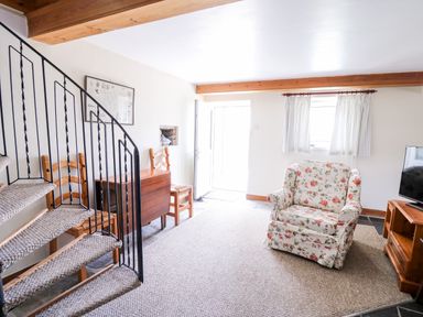 A living room with a floral armchair a wooden TV stand a TV a wooden table two chairs and a spiral staircase at Llety'r Wennol in Ffarmers near Lampeter