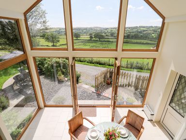 A dining area with a round table set with plates and flowers next to large windows overlooking a garden and countryside at Llety'r Wennol in Ffarmers near Lampeter