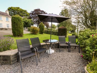 An outdoor seating area with four chairs a table and an umbrella on gravel beside a garden at Llety'r Wennol Ffarmers near Lampeter