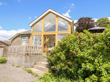Exterior of a stone house with large glass windows and wooden frames surrounded by bushes with flowers at Llety'r Wennol Ffarmers near Lampeter