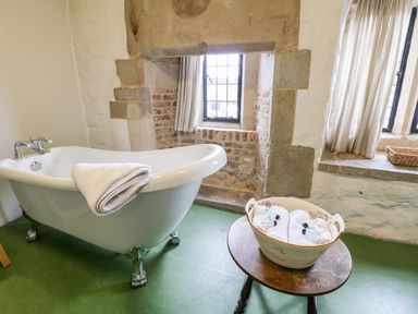A bathroom with a clawfoot bathtub a towel on the tub a small table with rolled towels and toiletries and windows with curtains at Porters Lodge in Polesworth