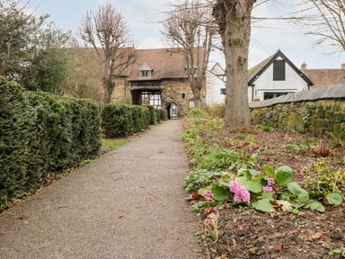 A pathway with bushes and flowers leading to a stone arch building and a white house at Porter's Lodge in Polesworth