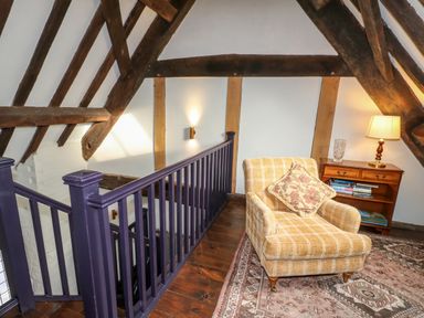 A sitting area with a yellow plaid armchair a wooden side table with books and a lamp near a staircase with purple railing in an attic space with exposed wooden beams and white walls at Gatehouse Croft in Polesworth