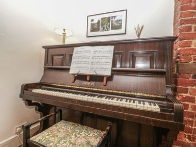 An upright piano with sheet music and a patterned bench in a corner with a brick wall and lamp at Church Farmhouse Winsham