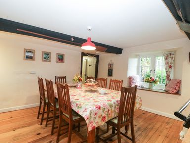 A dining room with a long wooden table covered by a floral tablecloth surrounded by wooden chairs and a window seat with cushions at Church Farmhouse in Winsham