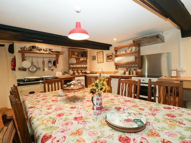 A kitchen with a floral tablecloth on a large wooden dining table with chairs and a flower vase at Church Farmhouse in Winsham