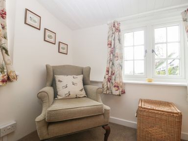 A corner of a room with an armchair with a duck-patterned cushion next to a window with floral curtains and a wicker basket at Church Farmhouse in Winsham