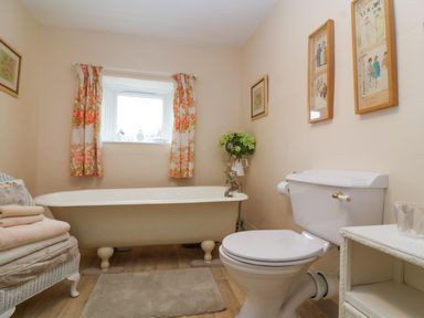 A bathroom with a bathtub under a window with floral curtains a toilet and a small table with framed pictures on the wall at Church Farmhouse in Winsham