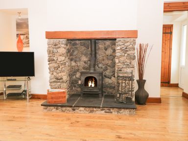 A stone fireplace with a wood burning stove burning fire a wooden box and fireplace tools on wooden floor at Fernleigh Cottage in Allithwaite near Grange over Sands