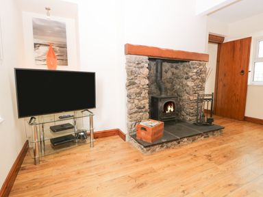 A room with a television on a glass stand and a stone fireplace with a wood burning stove at Fernleigh Cottage in Allithwaite near Grange-over-Sands