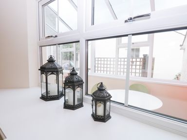 Three black metal lanterns with candles on a white window sill overlooking a white round table and green chairs at Fernleigh Cottage in Allithwaite near Grange-over-Sands