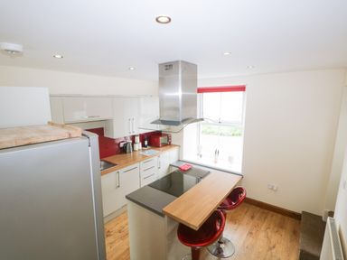 A kitchen with white cabinets red backsplash wooden countertop and two red stools at Fernleigh Cottage in Allithwaite near Grange-over-Sands
