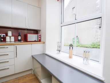 A kitchen corner with a red microwave on the countertop white cabinets wooden floor and utensils on the windowsill at Fernleigh Cottage in Allithwaite near Grange over Sands