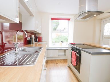 A kitchen with a stainless steel sink a stove with a red oven mitt and a window with a red blind at Fernleigh Cottage in Allithwaite near Grange-over-Sands