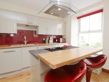 A kitchen with a wooden countertop red backsplash white cabinets and red stools at Fernleigh Cottage in Allithwaite near Grange-over-Sands