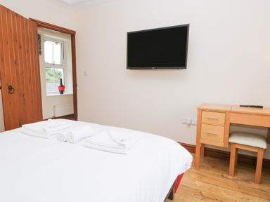 A bedroom with a bed, towels on the bed, a wooden door, a window with a vase, a wall-mounted TV, and a wooden desk with a stool at Fernleigh Cottage in Allithwaite near Grange-over-Sands