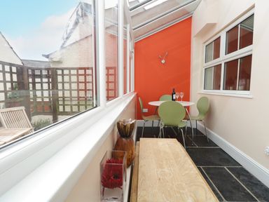 A narrow dining area with green chairs and a white round table against an orange wall with large windows at Fernleigh Cottage in Allithwaite near Grange-over-Sands