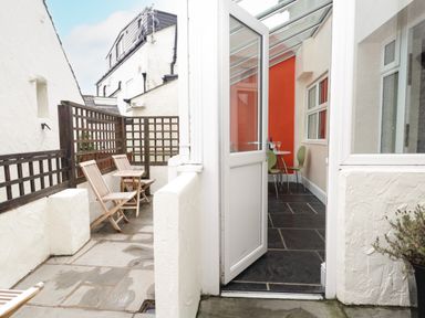 An outdoor patio with wooden chairs and a table next to a room with a glass door and green chairs at Fernleigh Cottage in Allithwaite near Grange-over-Sands