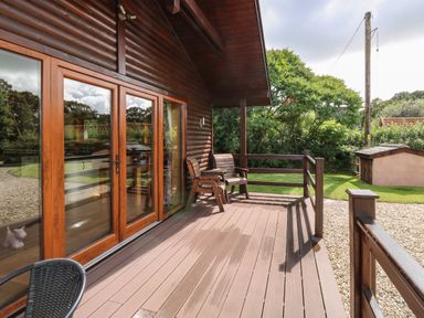 A wooden deck with two wooden chairs outside a house with large glass doors at Barn Shelley Lodge in Copplestone near Crediton