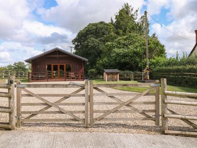A wooden gate leading to a gravel driveway with a wooden cabin and trees in the background at Barn Shelley Lodge in Copplestone near Crediton