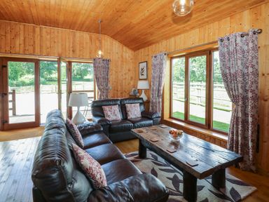 A living room with leather sofas a wooden coffee table floral curtains and large windows at Barn Shelley Lodge in Copplestone near Crediton