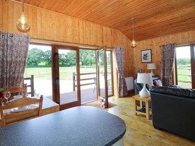 A wooden interior room with a dining table set, glass doors leading outside, black leather sofas, and lamps at Barn Shelley Lodge Copplestone near Crediton