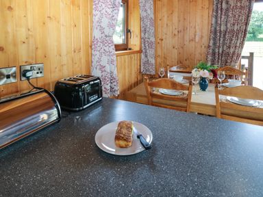 A kitchen countertop with a loaf of bread and knife next to a black toaster and a wooden dining table with chairs and place settings at Barn Shelley Lodge in Copplestone near Crediton